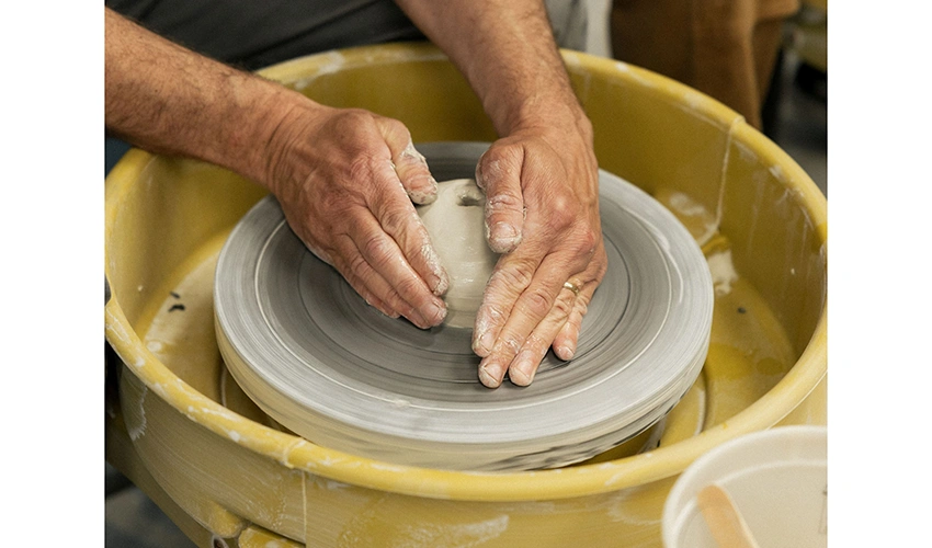 photo of a pair of lighter colored hands working clay in a yellow pottery wheel